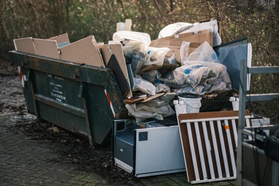 An overflowing dumpster with cardboard and bagged trash, and a discarded mini fridge and cabinet dumped on the ground beside it
